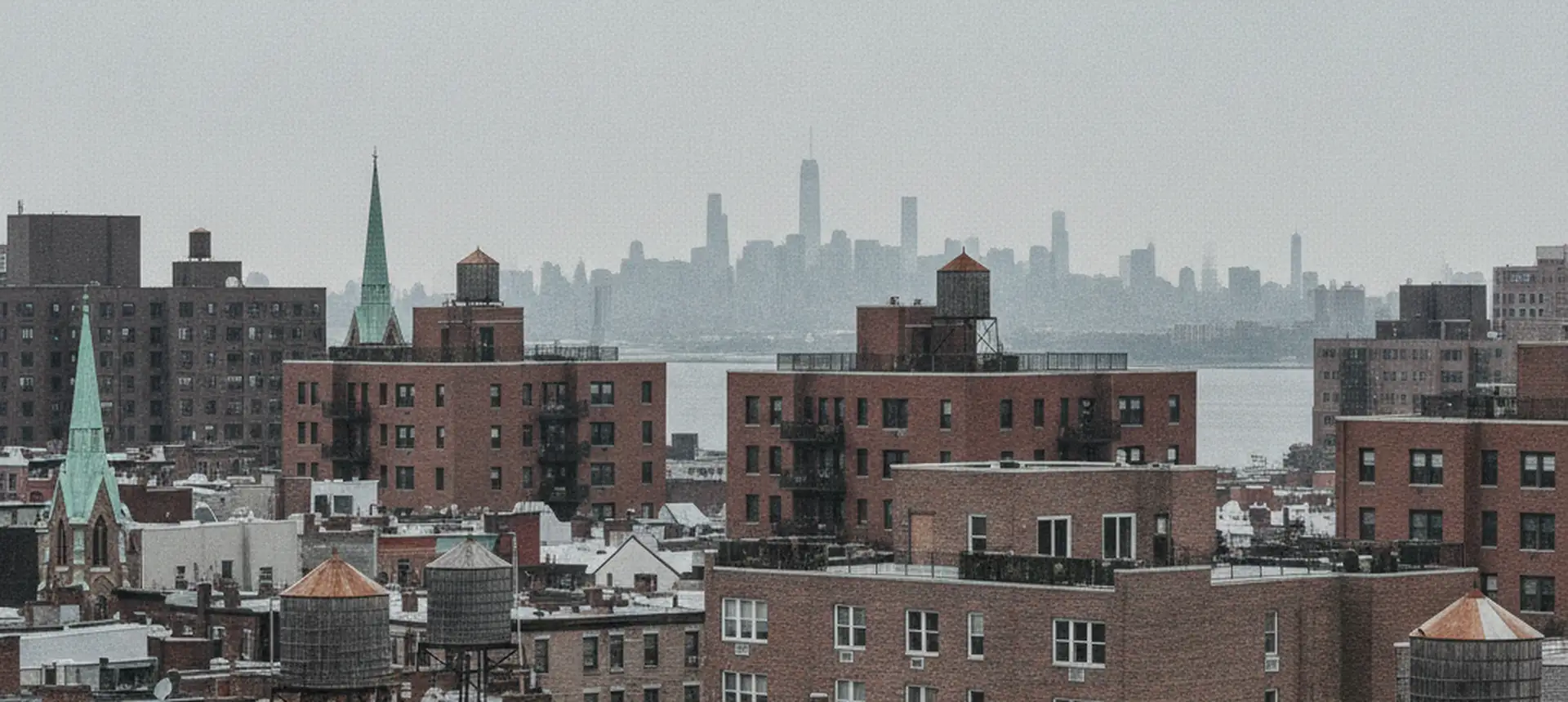 A wide view across Queens rooftops of low brick houses and mid-rise apartment buildings toward a distant skyline, water towers and church spires.