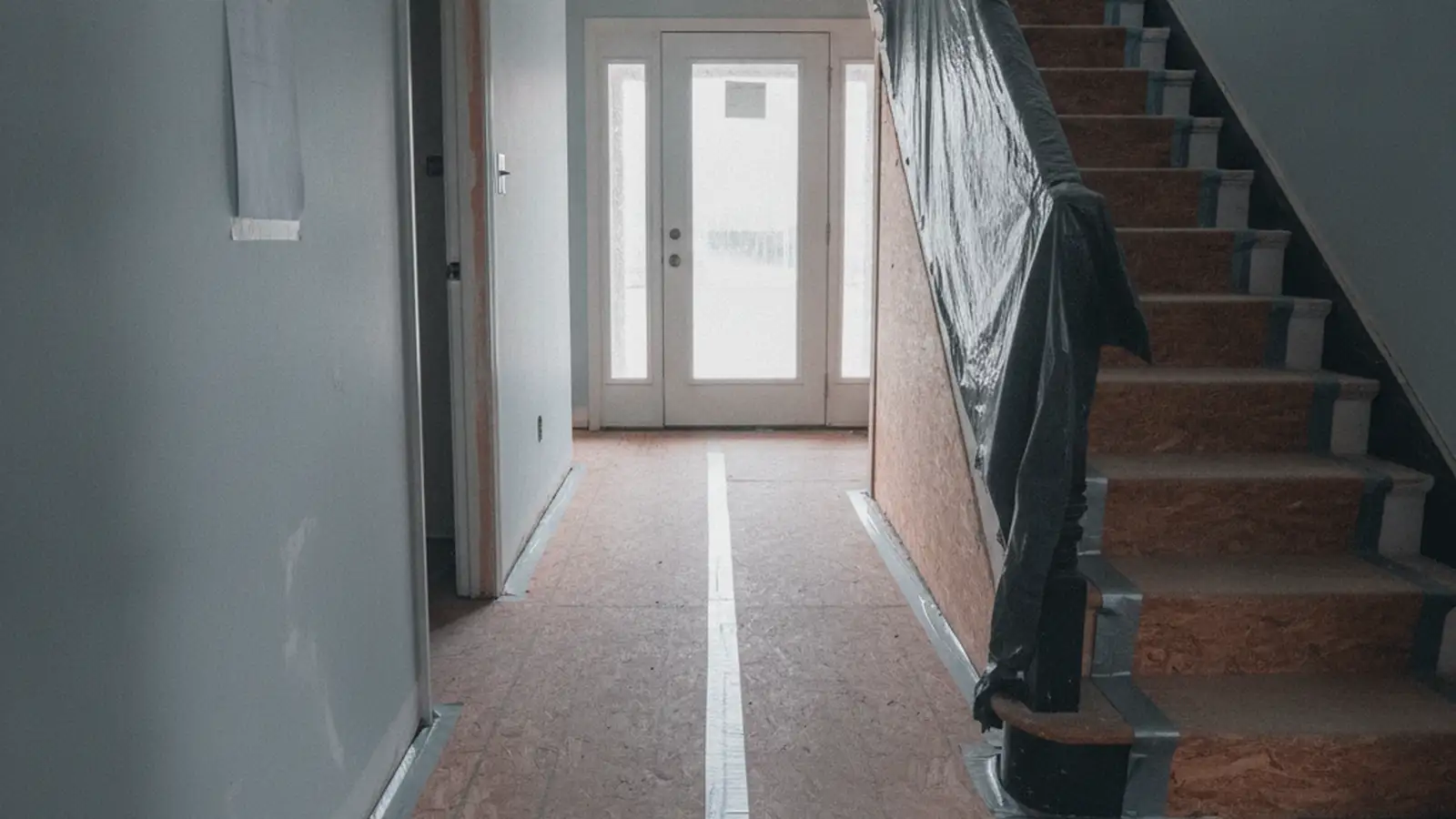 The front hall of a Queens two-family house with the tile floor and the stair covered in protective board, daylight through the door glass.